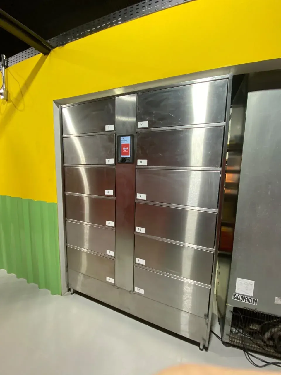 Stainless steel locker with 12 numbered doors and the kiosk in the middle, set into a restaurant’s yellow wall